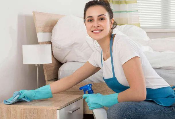 A representative of expert cleaning services is demonstrating the art of deep cleaning while cleaning the side table next to the bed in the room.