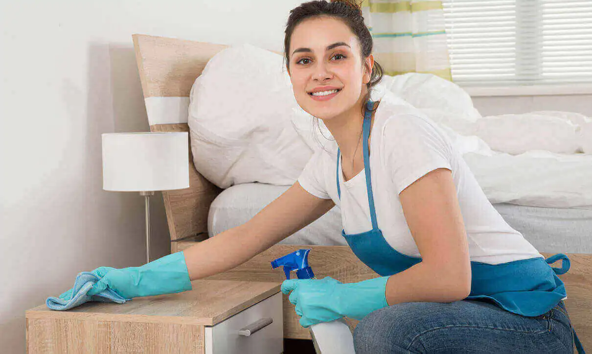 A representative of expert cleaning services is demonstrating the art of deep cleaning while cleaning the side table next to the bed in the room.